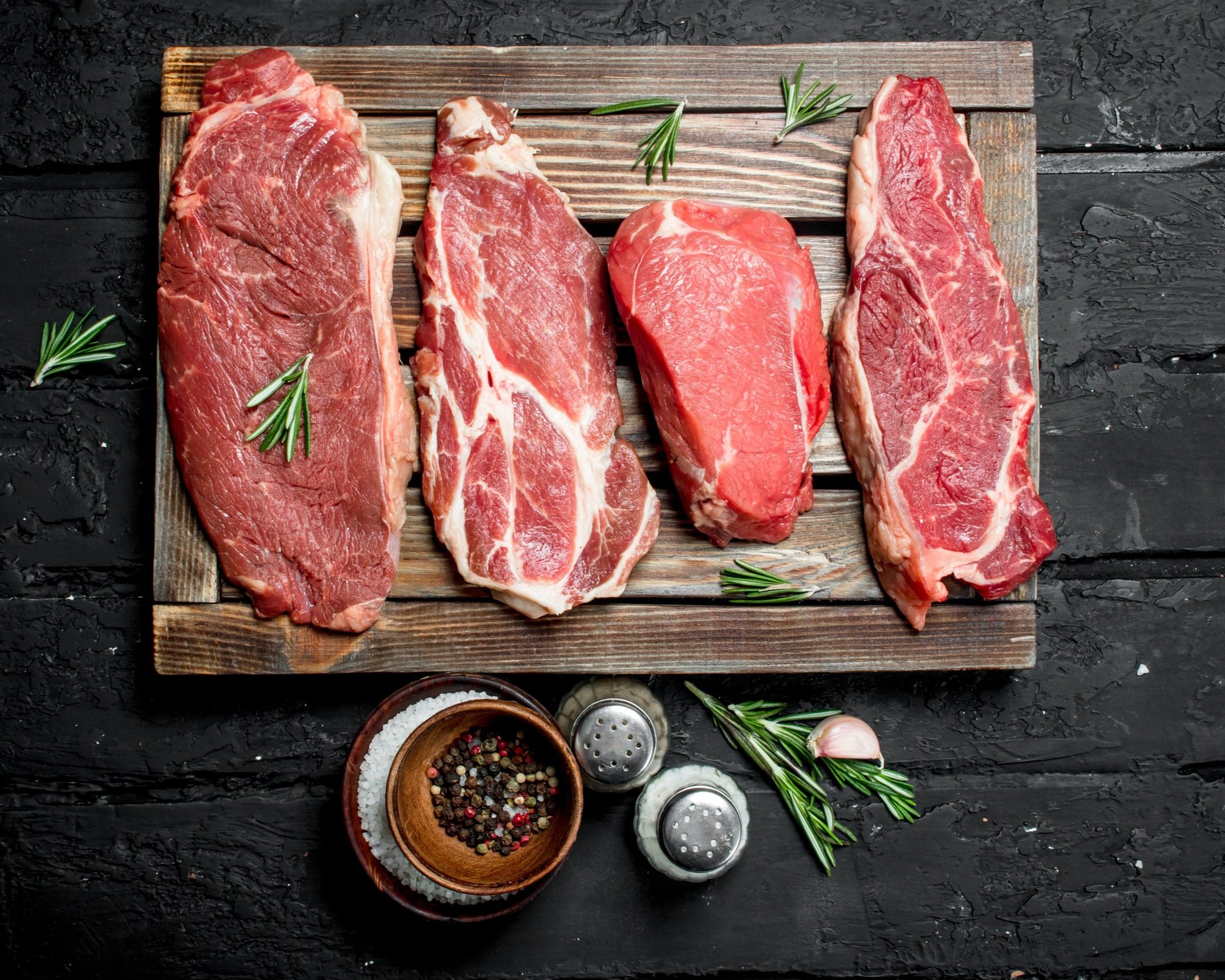 Assorted cuts of raw beef steaks displayed on a wooden board, garnished with fresh rosemary, accompanied by a bowl of mixed peppercorns, salt, and garlic on a dark textured surface.