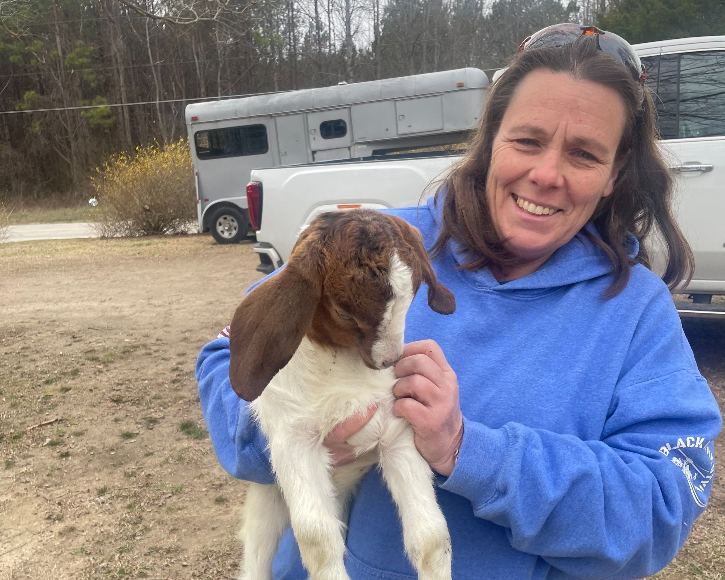 Misty+hawk+-+9FB51E8D-A946-4706-817F-5E0F6ACA71ED Woman smiling while holding a brown and white goat in a rural setting, with a horse trailer and pickup truck in the background.