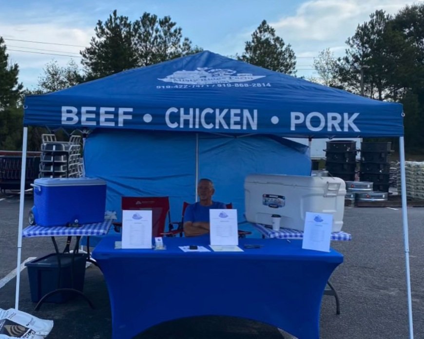 Misty+hawk+-+D4EAB123-BD5A-444C-A56F-BEB8F0AA2E7E A vendor at a food stand under a blue canopy displaying "BEEF • CHICKEN • PORK," with a cooler and menu on the table, set in a parking lot surrounded by trees and equipment.