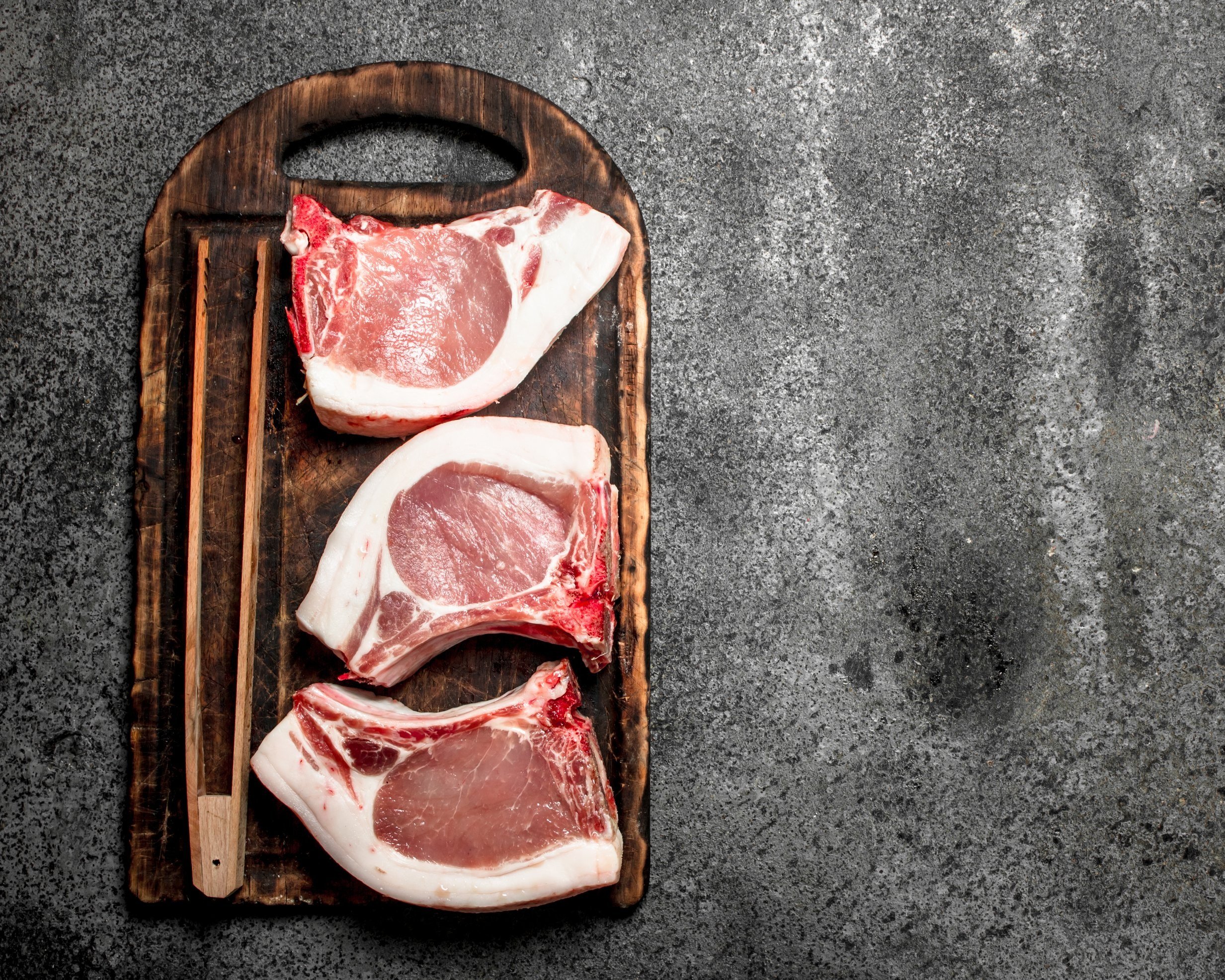 Fresh pork chops displayed on a rustic wooden cutting board, accompanied by chopsticks, set against a textured gray background. Ideal for recipes and cooking inspiration.